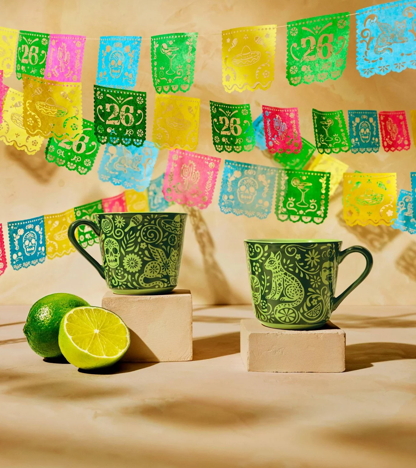 Två gröna muggar med intrikata mönster, en hel och en halv lime, under färgglada papel picado-banderoller.