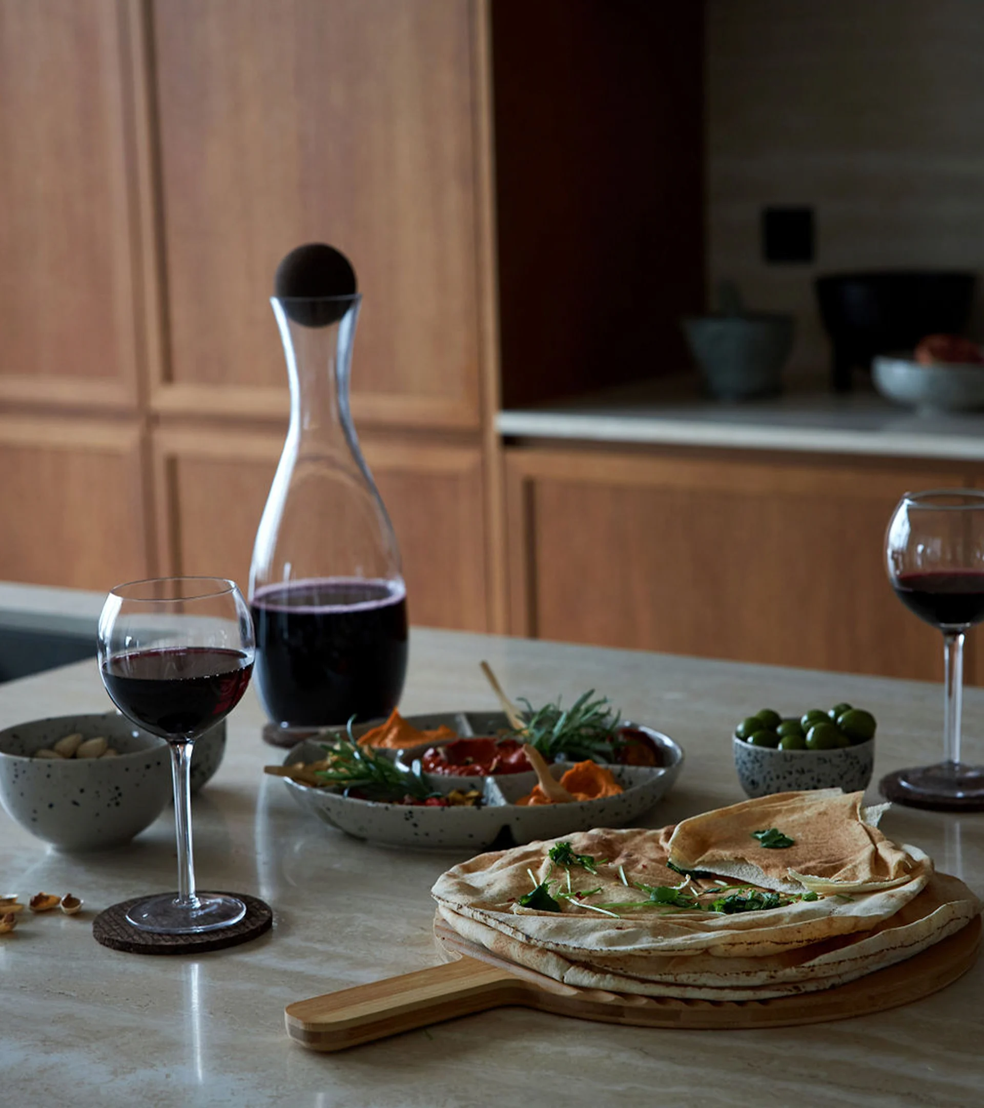 Ett bord dukat med rödvin, flatbröd, dippar, oliver och nötter, mot en varm köksbakgrund i trä.