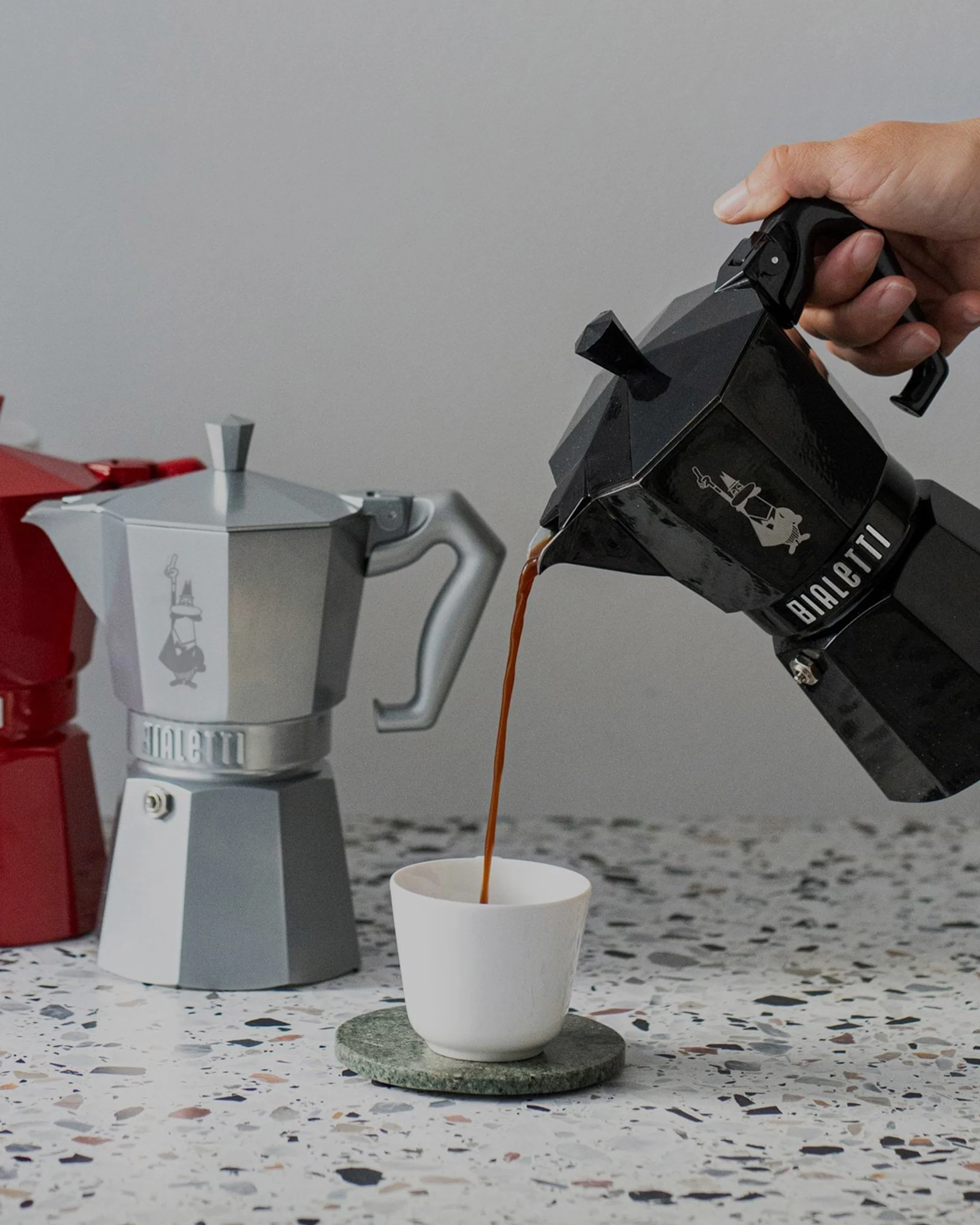 Hand häller kaffe från svart mokkabryggare i vit kopp. Silver och röd mokkabryggare i bakgrunden.