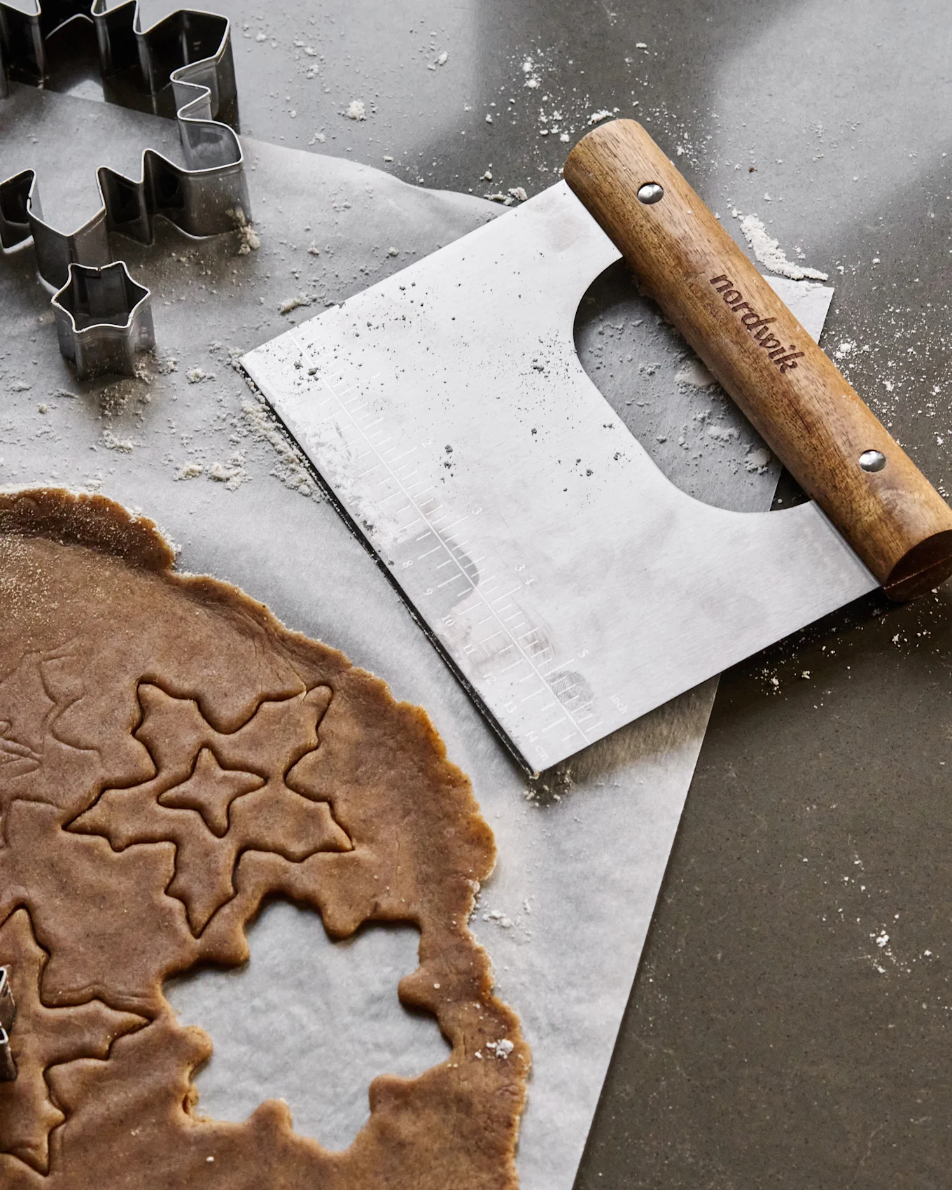 Pepparkaksdeg med stjärn- och lövformar, metallkakformar och en degskrapa på en mjölad yta.