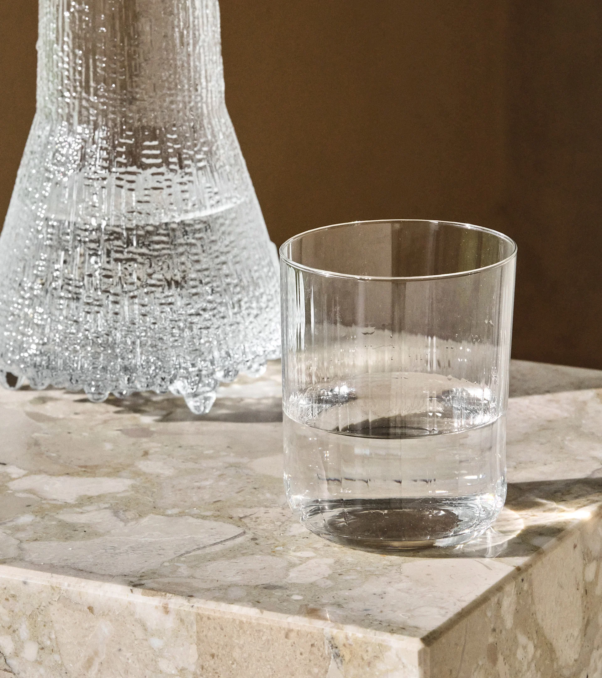 En klar, texturerad glaskaraff och ett slätt dricksglas, båda med vatten, står på en ljus marmoryta.