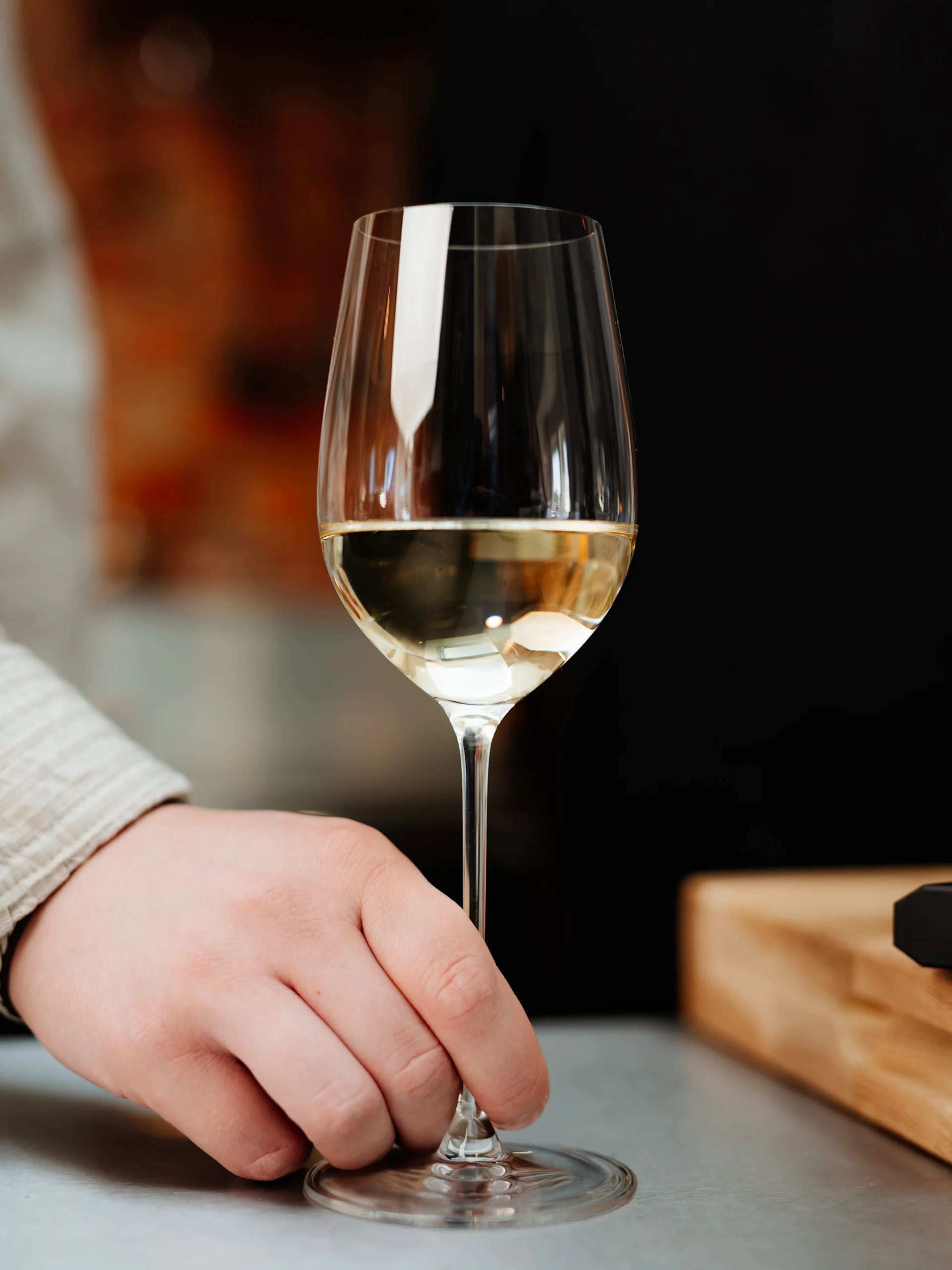 En persons hand håller i stjälken på ett vinglas fyllt med vitt vin på en ljus yta.