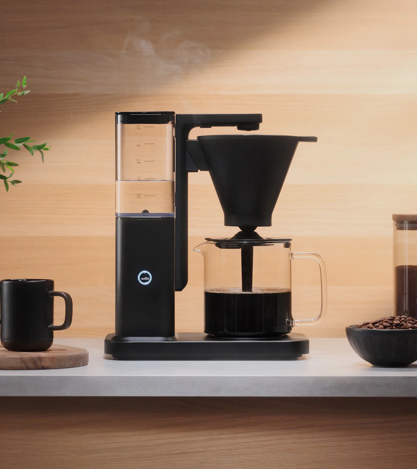 En svart kaffebryggare brygger kaffe i en glaskanna på en ljus bänkskiva, med ånga som stiger. En svart mugg och kaffebönor finns i närheten.