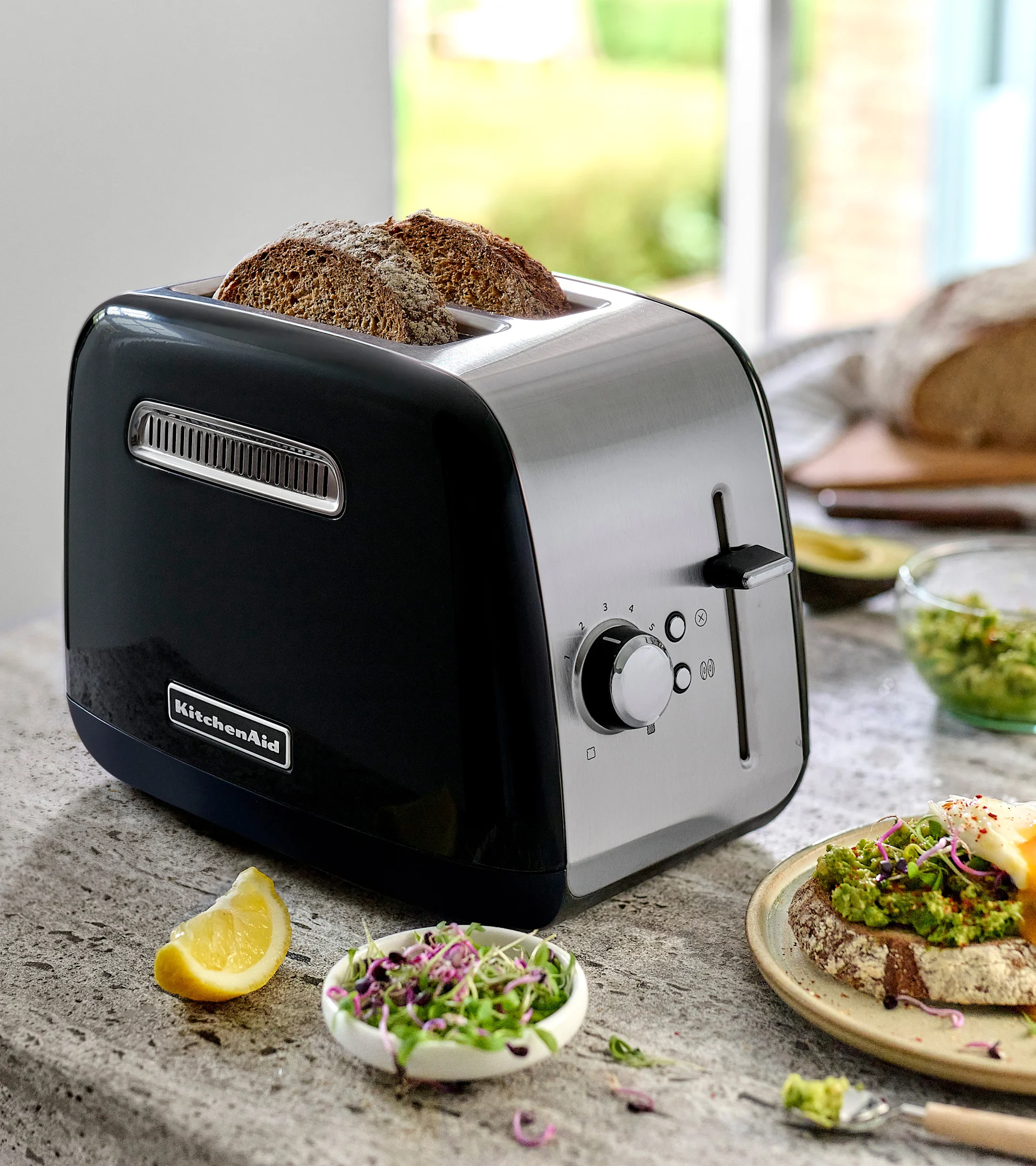 En svart och rostfri KitchenAid brödrost med två skivor mörkt bröd. På bänken, avokadotoast med ägg, groddar och en citronklyfta.