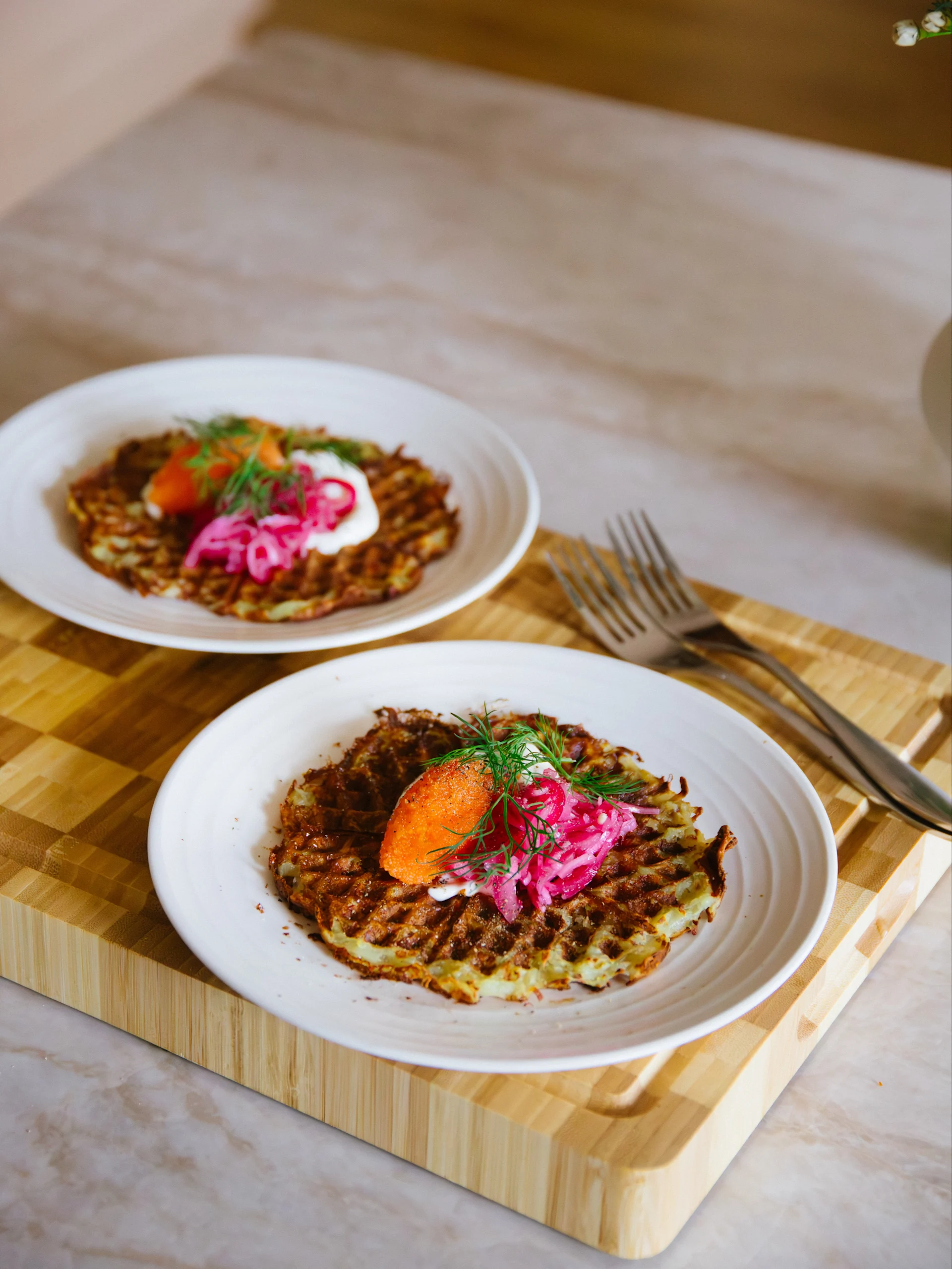 Två tallrikar med krispiga potatisvåfflor toppade med orange rom, gräddfil, inlagd rödlök och dill på en träskärbräda.