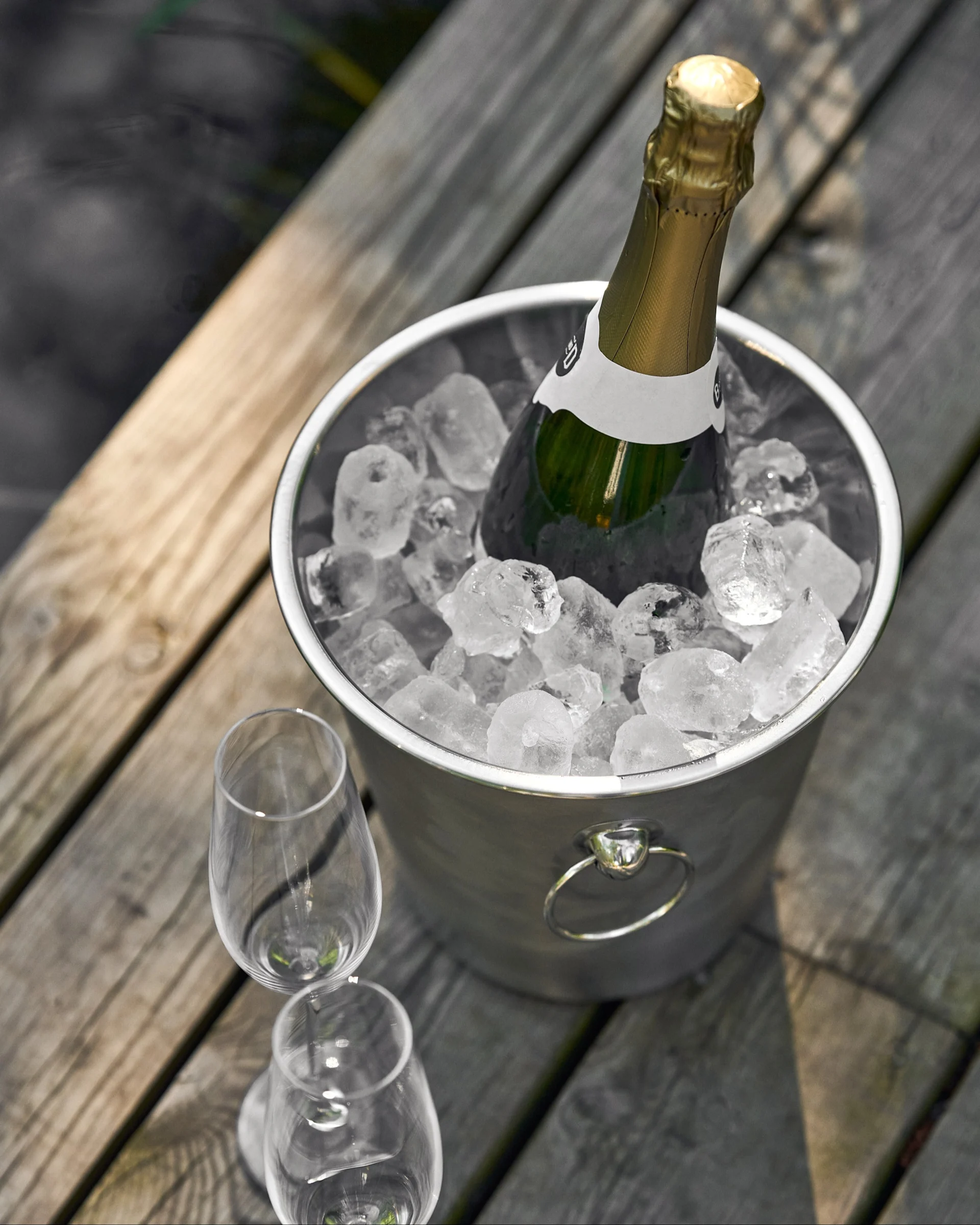 Champagneflaska i en ishink med två glas på ett trädäck vid vattnet.
