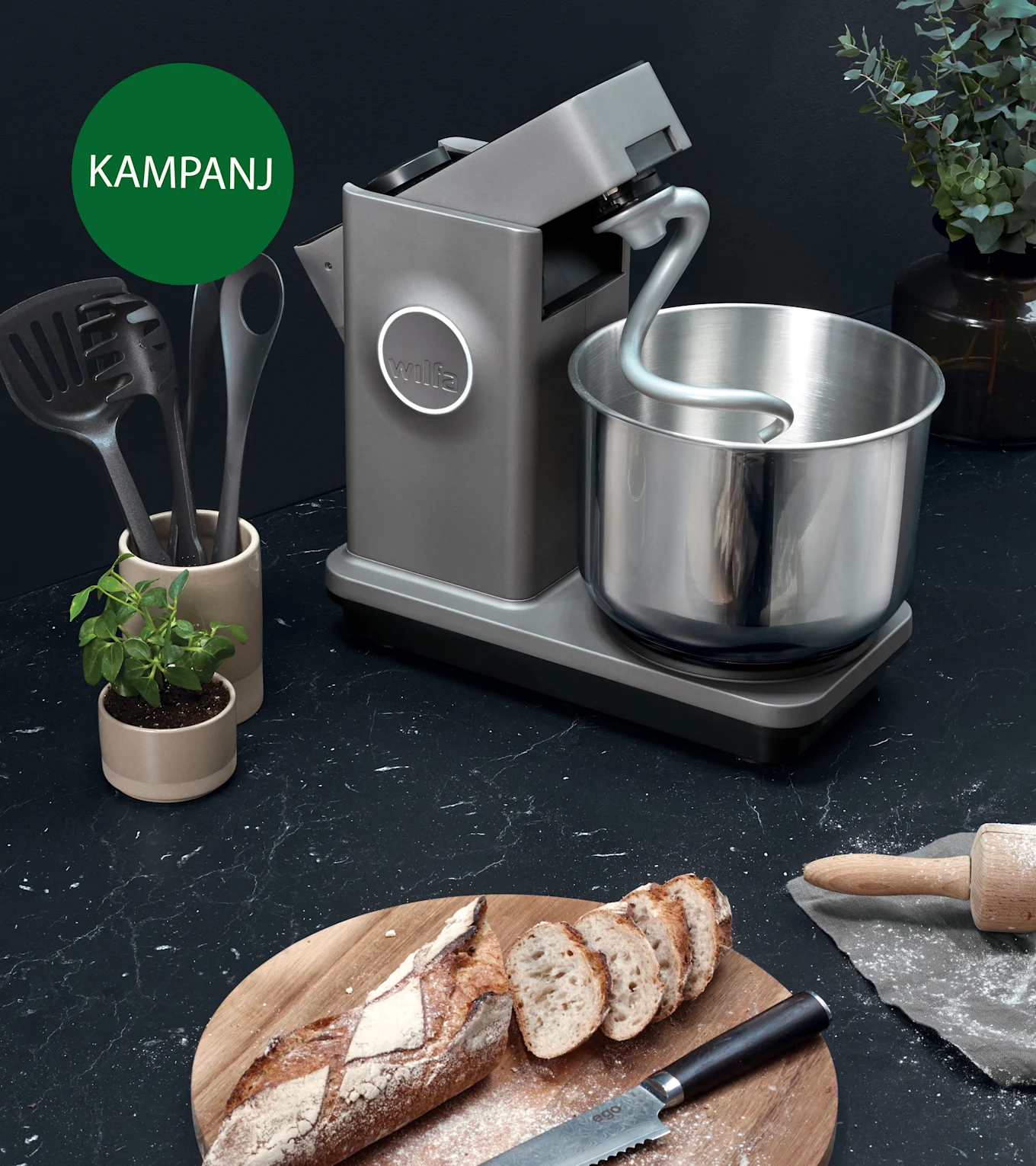 Grå köksmaskin med degkrok och stålskål på en mörk köksbänk med bakat bröd. En grön kampanjcirkel i övre vänstra hörnet.