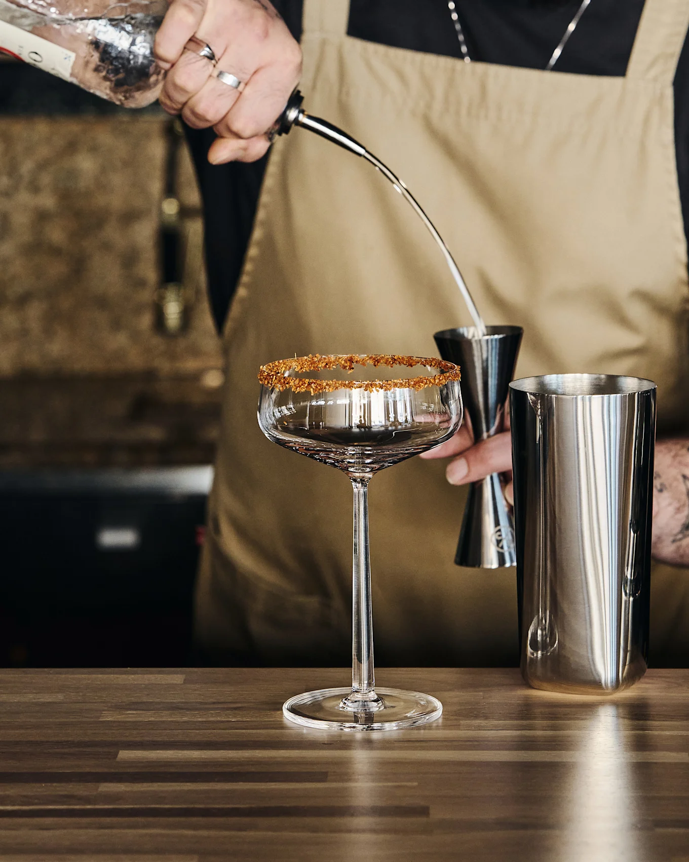 Bartender häller vätska från en flaska i ett mätglas över en shaker, bredvid ett glas med sockerkant.