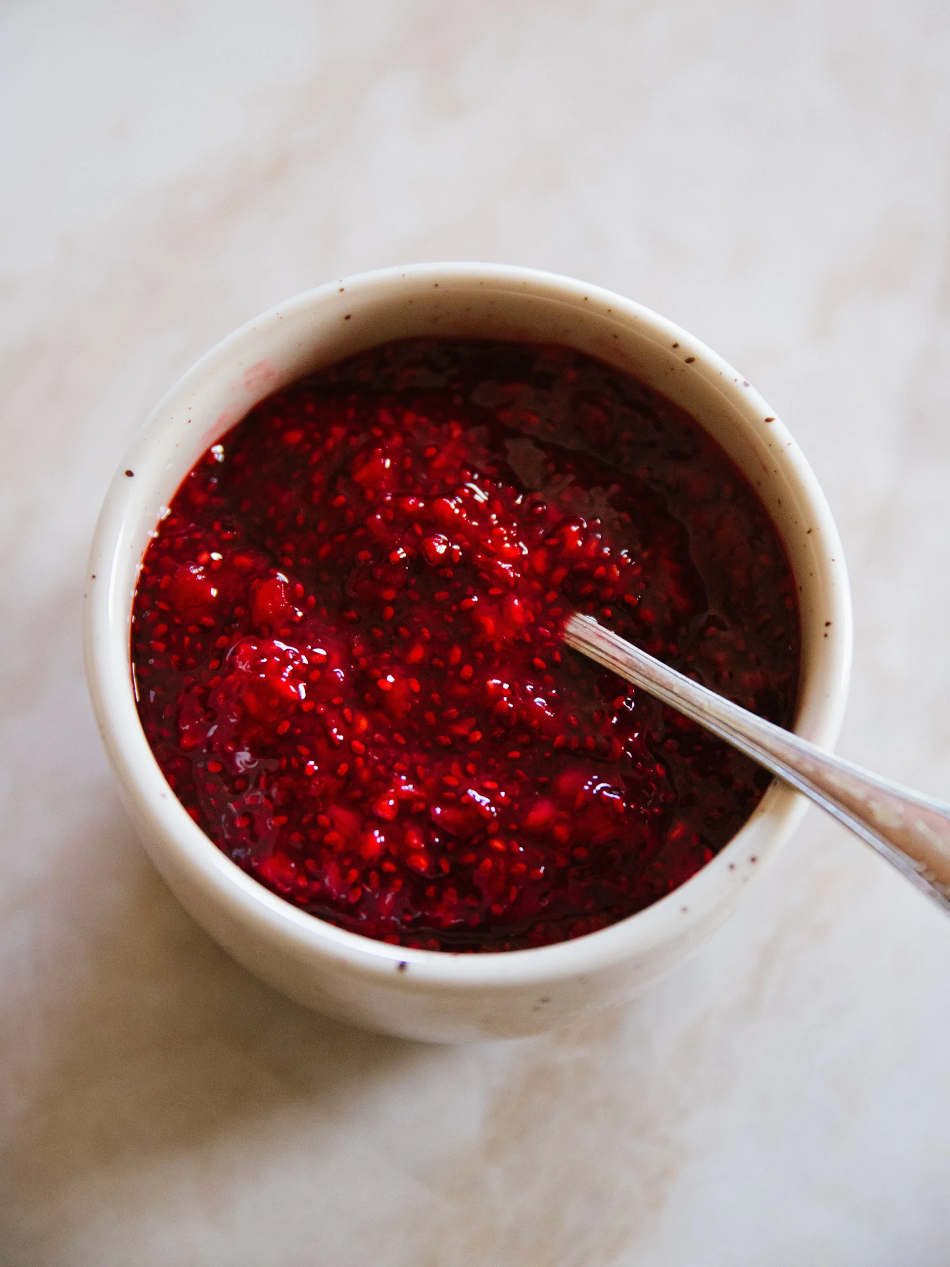 En vit skål fylld med livfullt röd hallon-chia-pudding med en silversked i, på en ljus bakgrund.