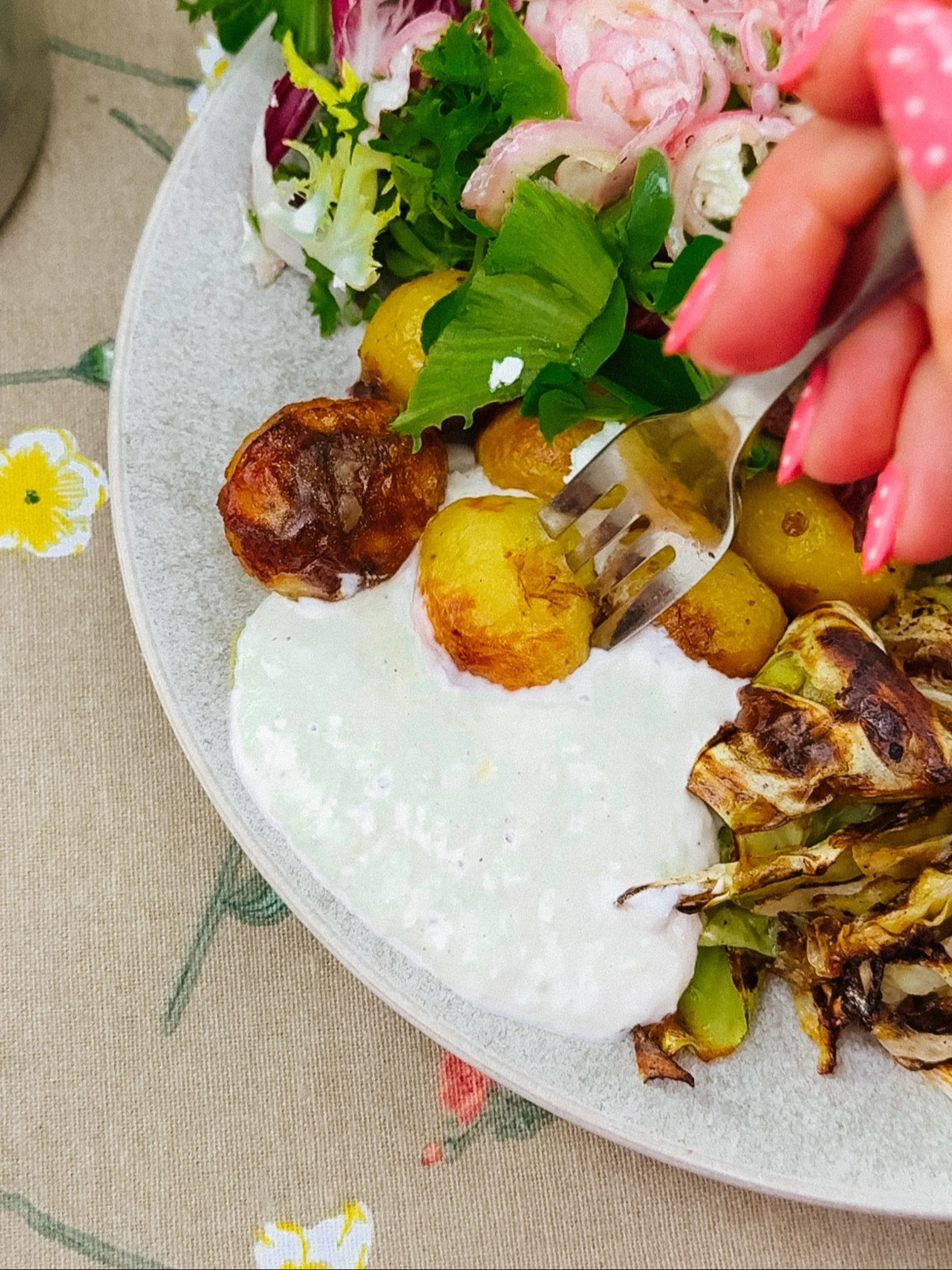 En hand med rosa naglar tar gyllene rostade potatisar och krämig sås från en tallrik med sallad och rostad kål.