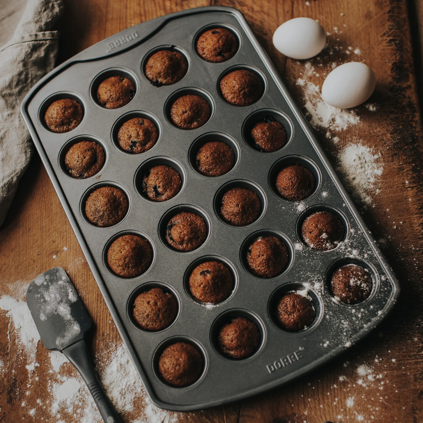 En muffinsform med 24 bakade minimuffins på en träyta med mjöl, två vita ägg och en spatel.