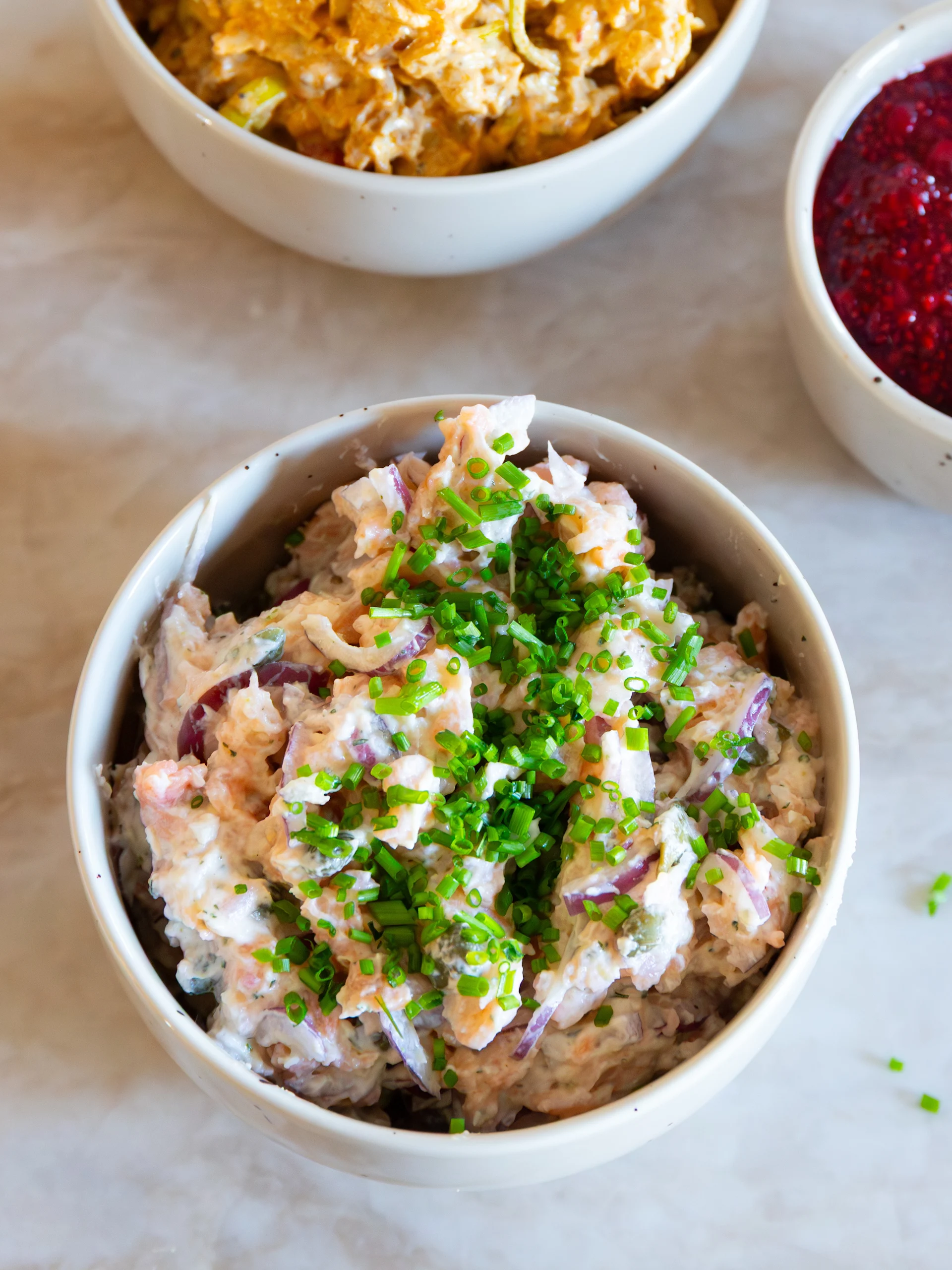 Krämig rosa sallad med rödlök och grön gräslök, med suddiga skålar med gul sallad och röd bärsylt i bakgrunden.