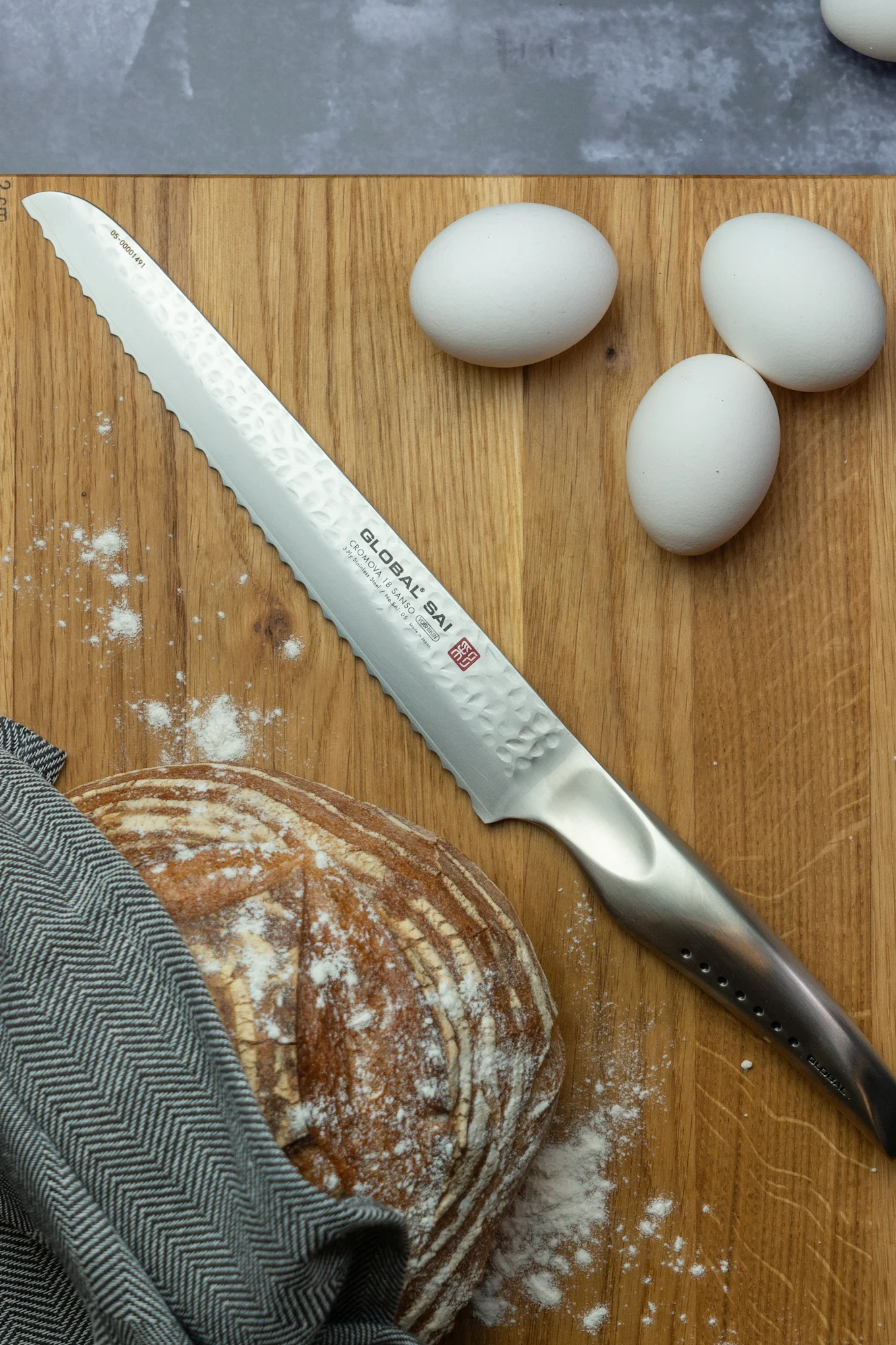 Global Sai SAI-05 brödkniv som tillhör Global knivserien SAI, ligger på ett träbräde med bröd och ägg.