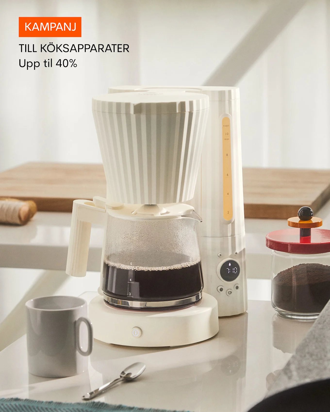 En vit kaffebryggare med en glaskanna fylld med kaffe, bredvid en grå mugg och en burk med kaffepulver. Text: Kampanj till köksapparater, upp till 40% rabatt.