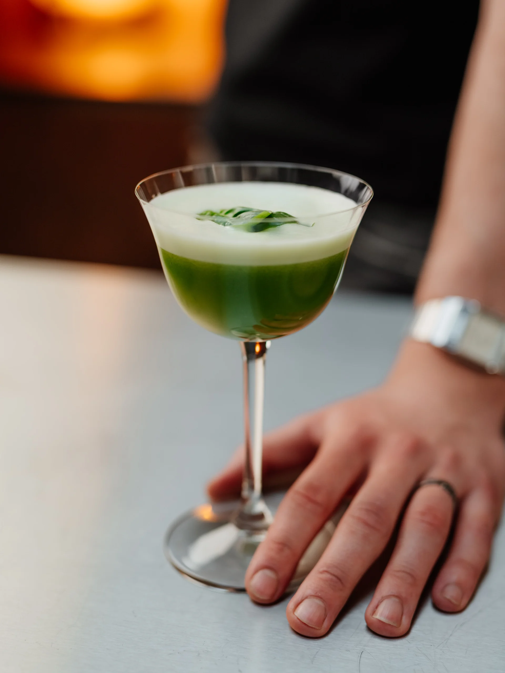 En grön och vit skiktad cocktail med blad i ett coupeglas, hållen av en hand med en ring.