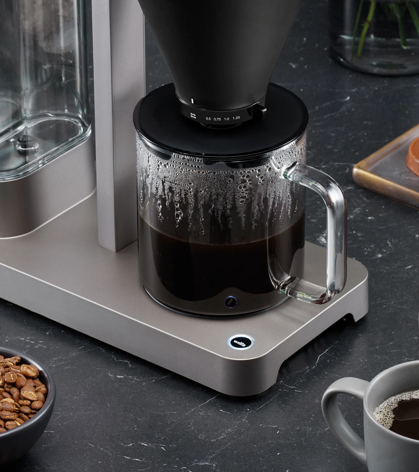 En modern kaffebryggare med svart filterkon brygger kaffe i en glaskanna på mörk bänkskiva, med kaffebönor och en mugg.