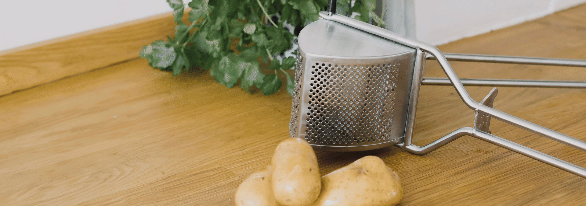 En potatispress i metall bredvid flera råa potatisar på en träbänk, med gröna örter i bakgrunden.