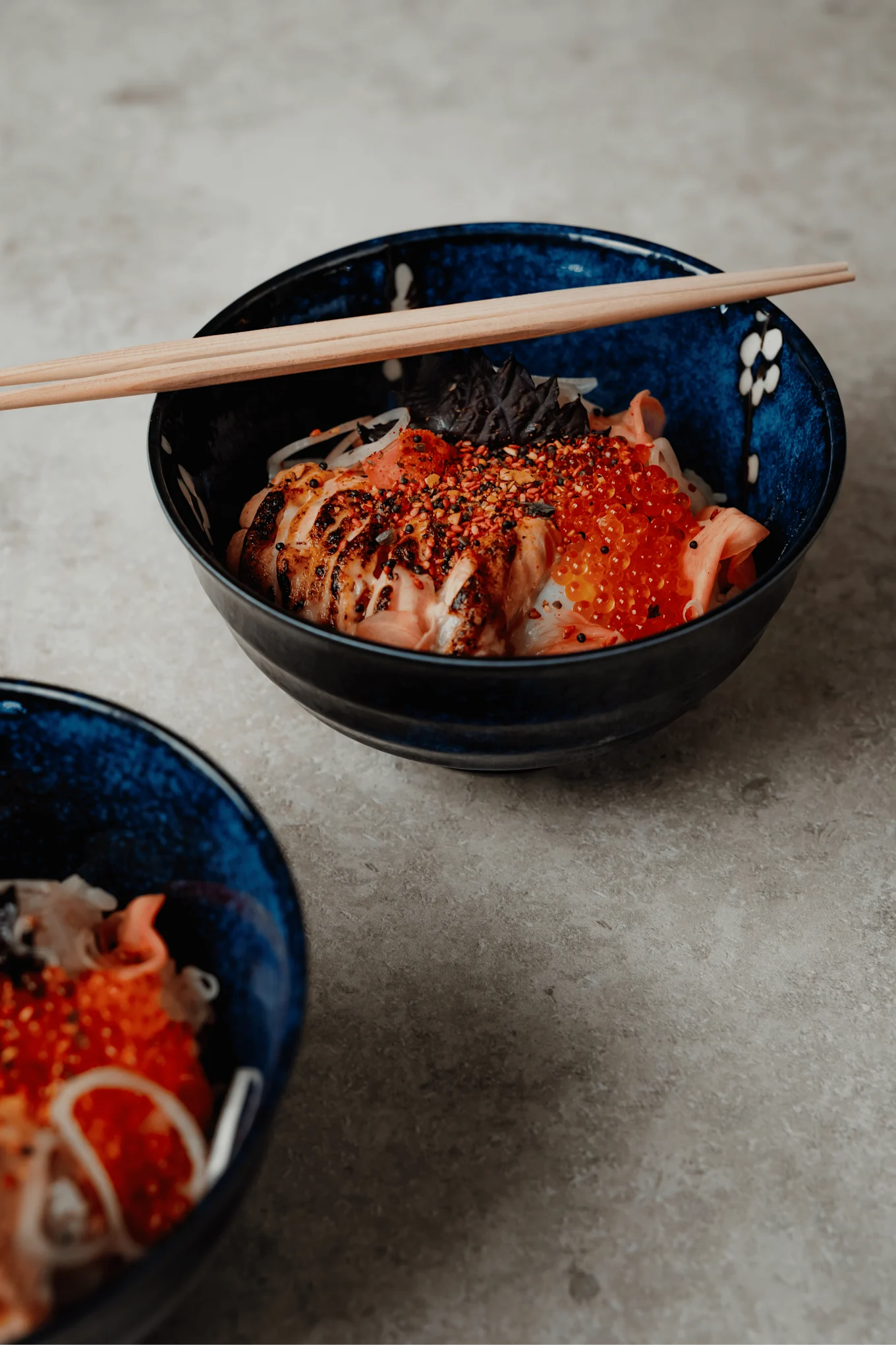 Två mörkblå skålar med japansk mat. Främre skålen visar grillad lax, laxrom, lök, kryddor och ätpinnar.