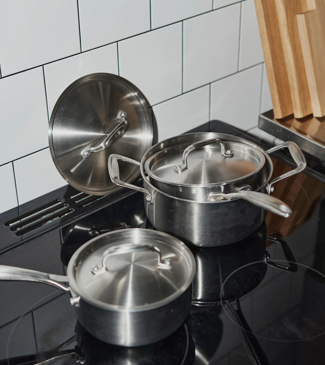 Grytor och lock i rostfritt stål på en svart induktionshäll, med vitkaklat stänkskydd och träskärbrädor.