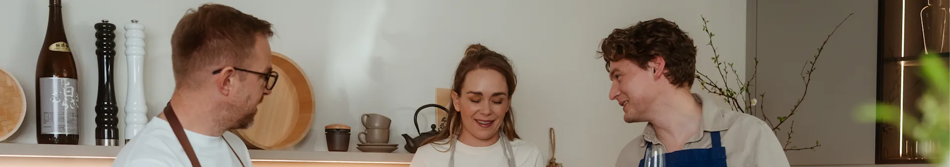 Tre personer, två män och en kvinna, bär förkläden och ler medan de lagar mat i ett modernt kök.