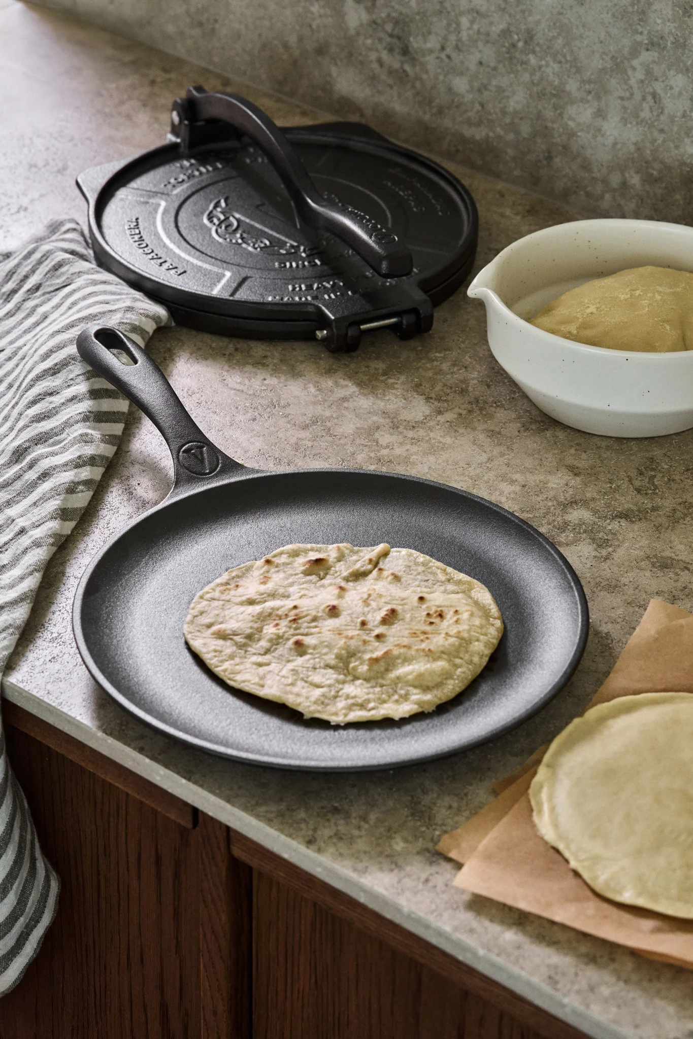 En gjutjärnspanna på köksbänken med en stekt tortilla, bredvid den står en gjutjärnspress för tortillor och en skål med deg. 