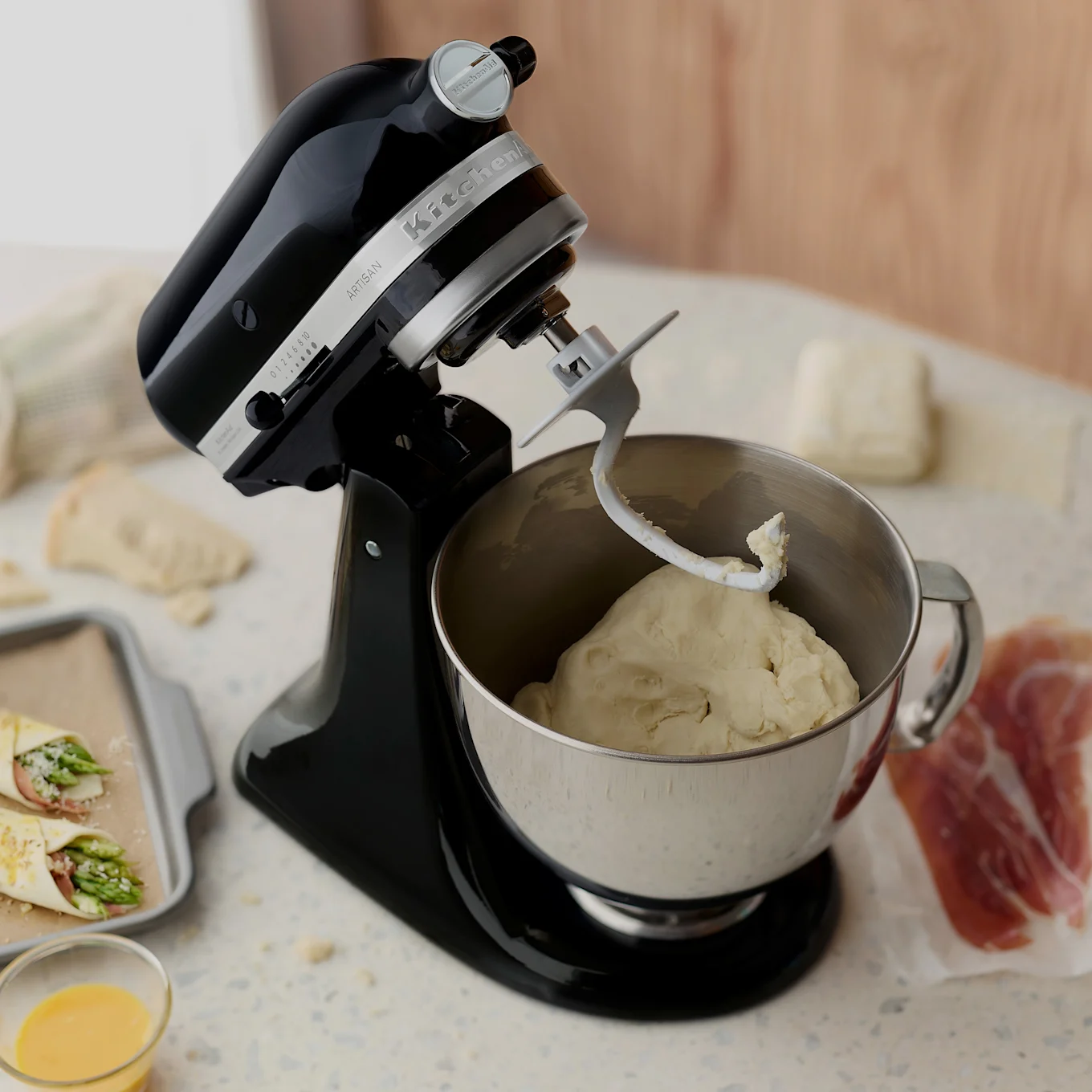 Svart köksmaskin med degkrok i en degskål av rostfritt stål, omgiven av ingredienser för salta bakverk.