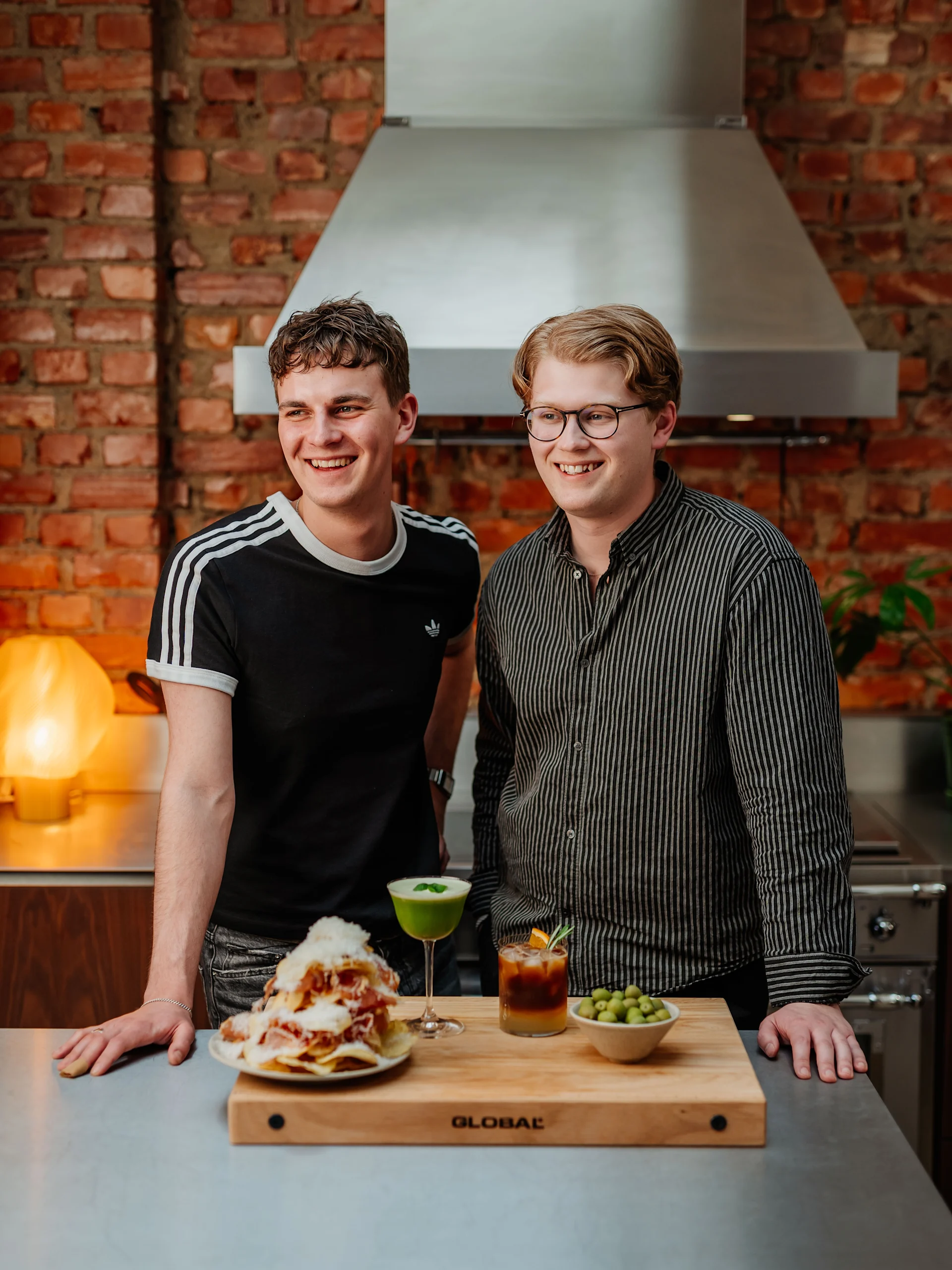 Två leende män bakom en disk med nachos, cocktails och oliver.