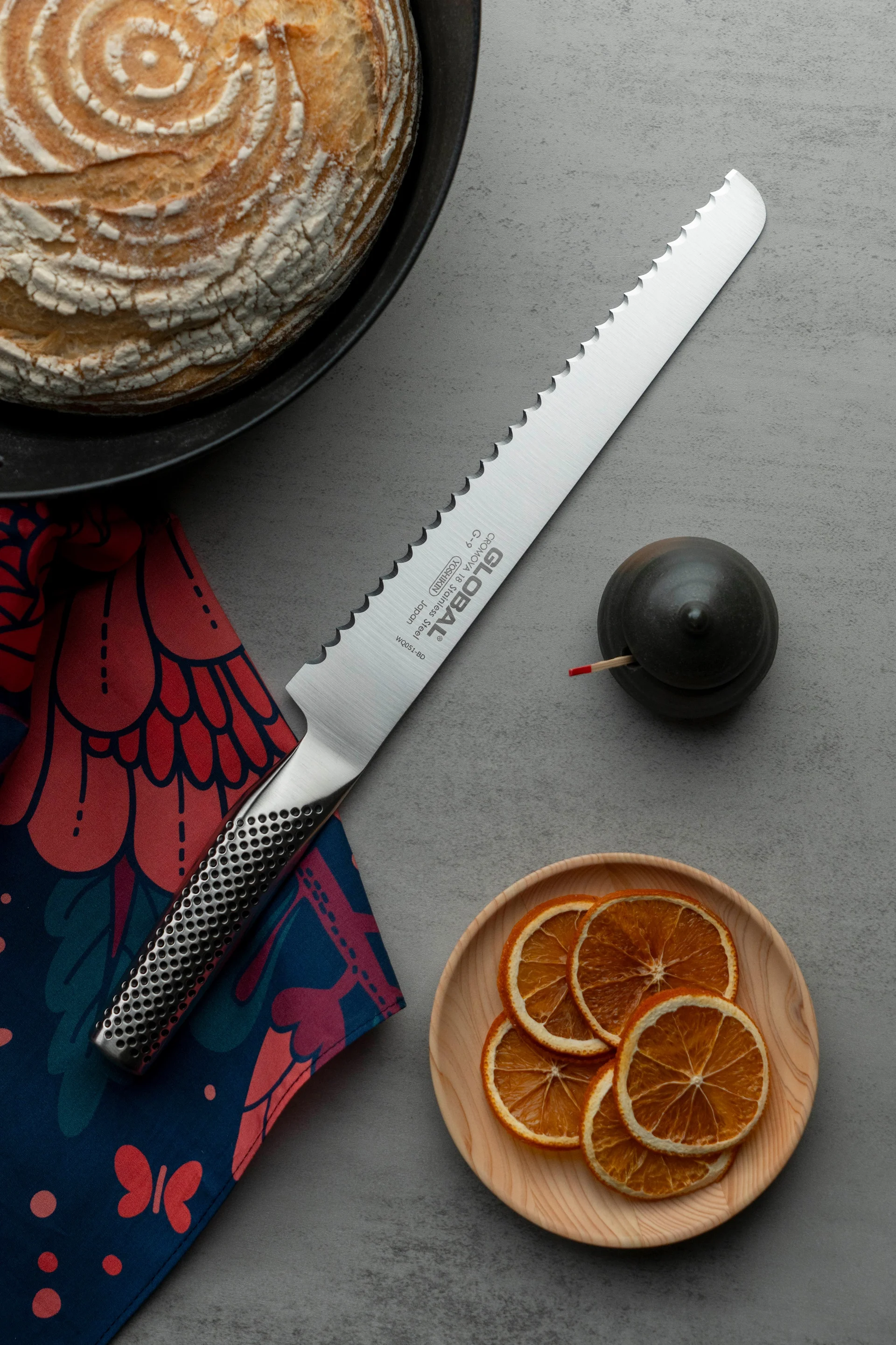 Global G-9R brödkniv syns tillsammans med ett bröd, torkade apelsiner och en handduk.