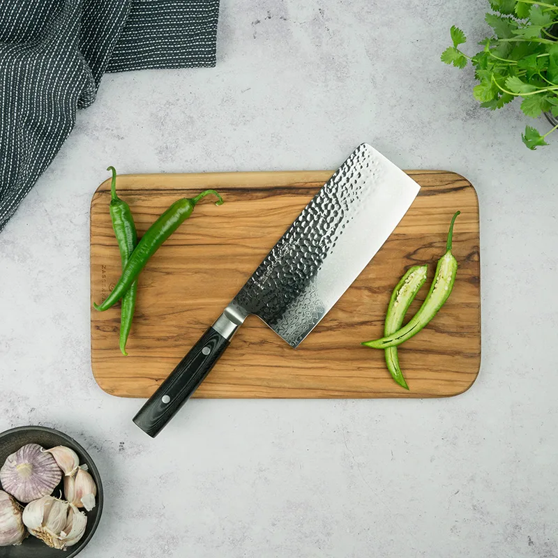 Nakiri-kniv med hamrat blad på en skärbräda i trä med gröna chilifrukter och vitlök.