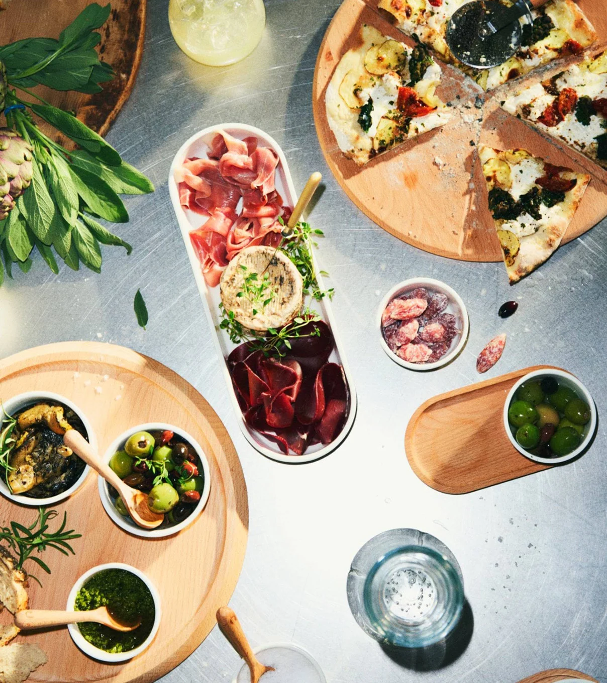 Ovanifrån bild av en matuppdukning med skivad pizza, charkbricka, oliver, pesto och tapenade på träbrickor.