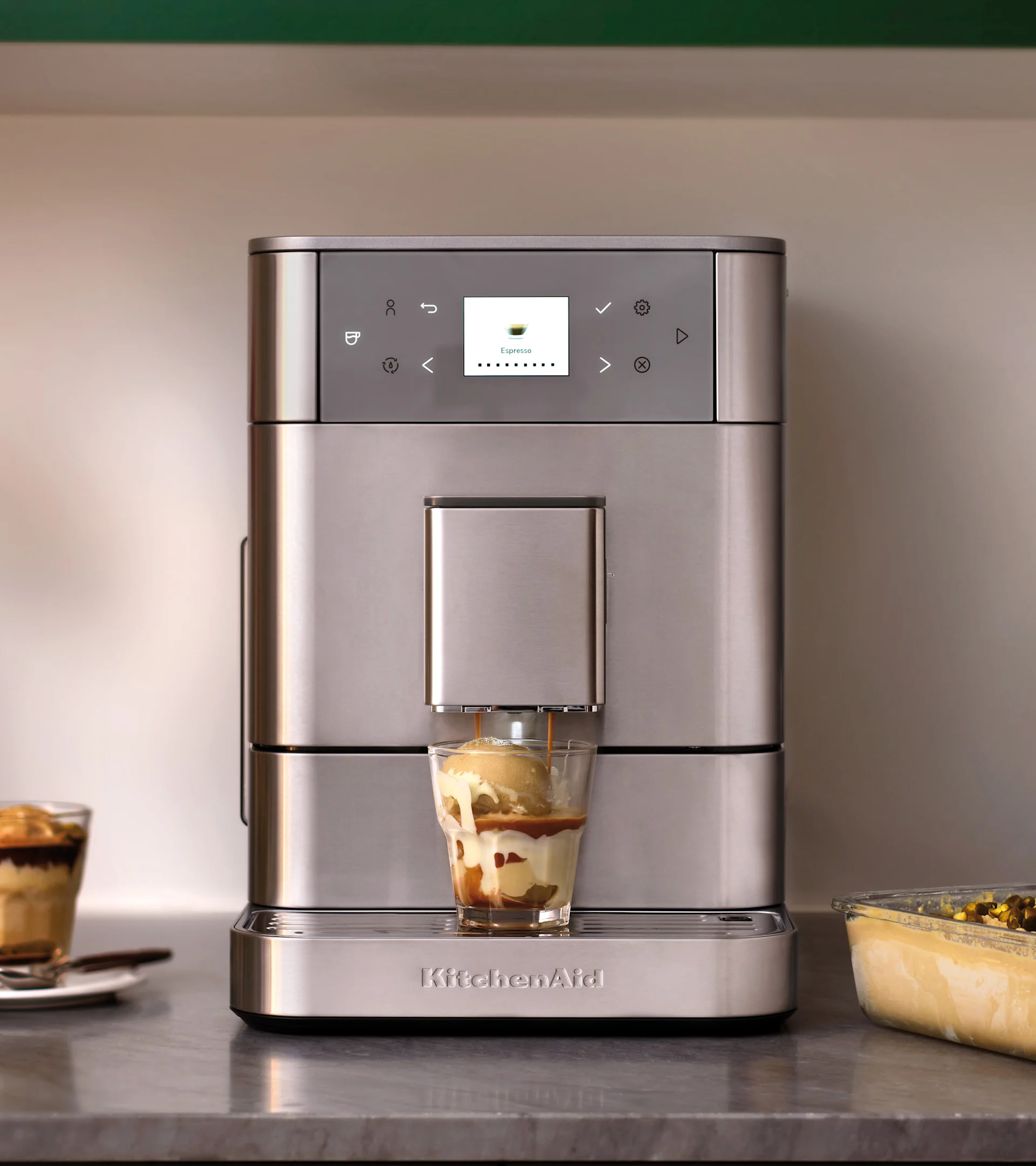Silverfärgad KitchenAid espressomaskin häller espresso i ett glas glass, gör en affogato. Annan dessert i bakgrunden