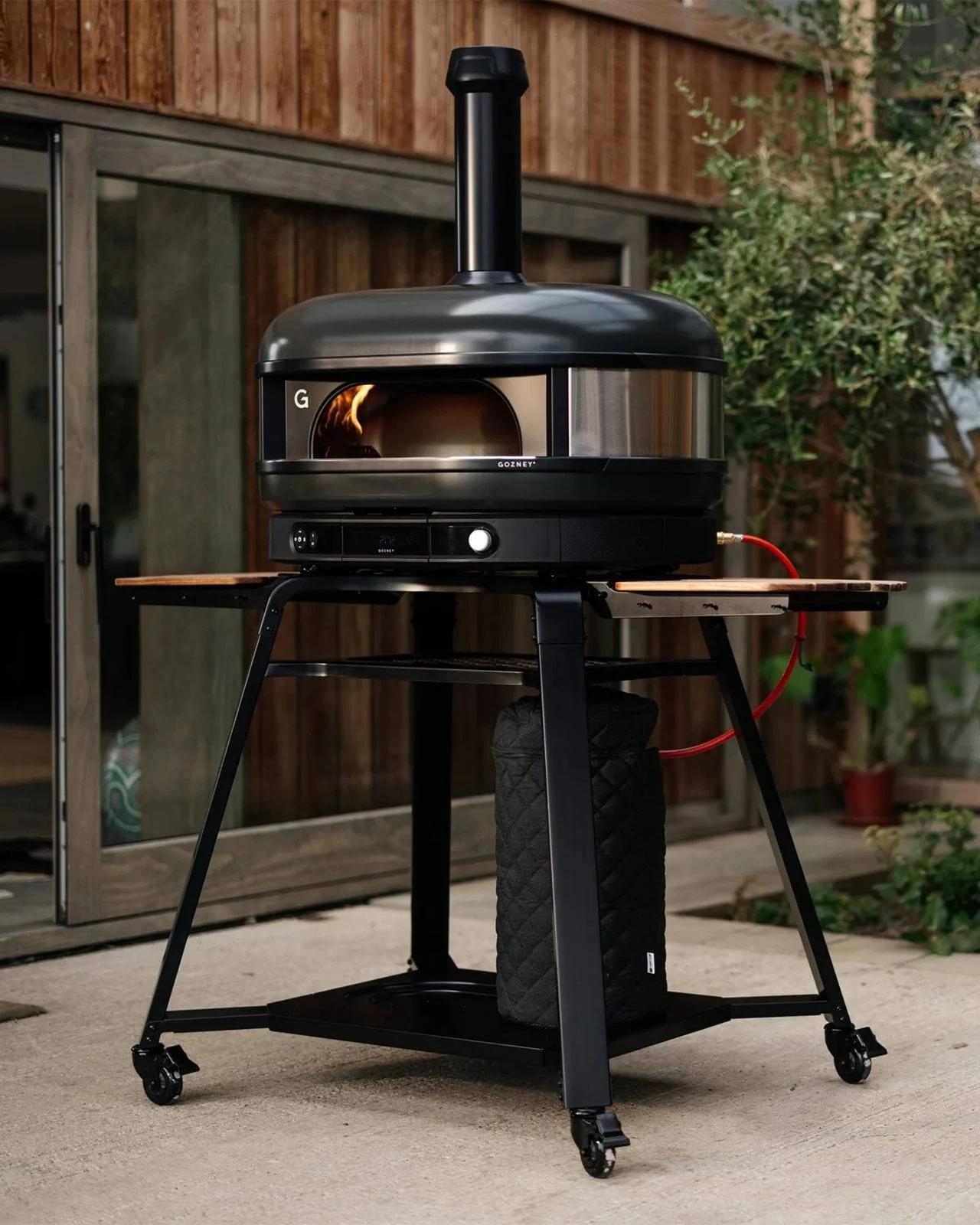 Svart utomhuspizzaung med synlig flamma, på ett rullbart stativ med en trähylla och en övertäckt gastank.