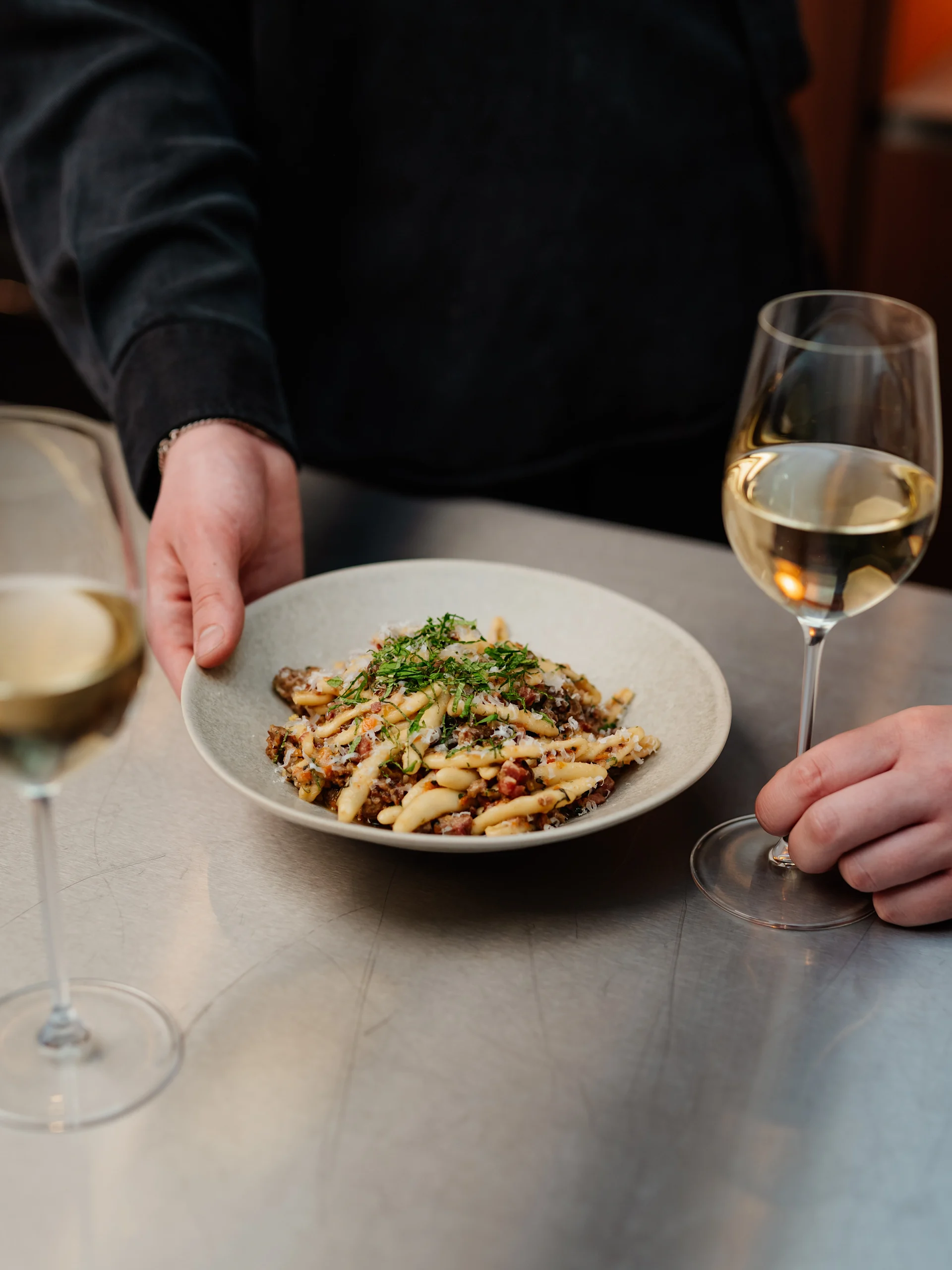 En persons händer håller en tallrik pasta med köttfärssås, ost och örter, som placeras på ett metallbord bredvid en annan persons hand som håller ett glas vitt vin. Ett andra vinglas är i förgrunden.