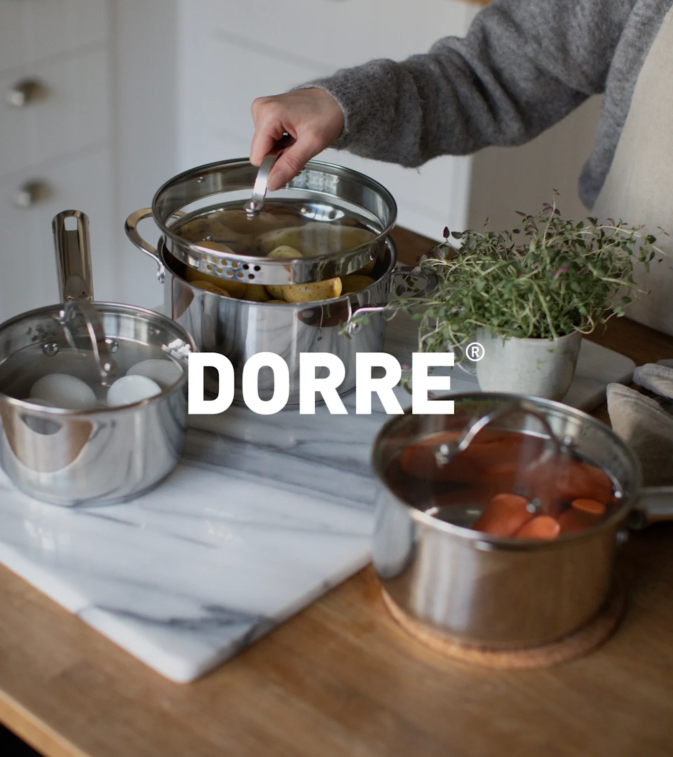 Hand lyfter lock från ångkokare i rostfritt stål med potatis, bredvid kastruller med ägg och morötter. DORRE-logotyp.